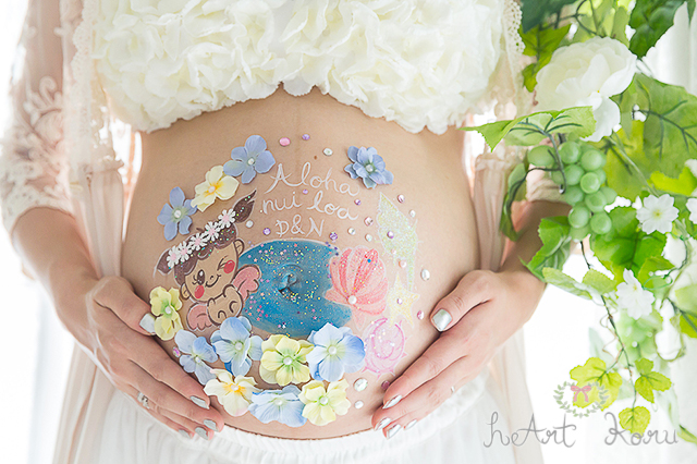 お姫様コースのベリーペイントのデザイン例。黄色とブルーのお花で彩られているとてもおしゃれなマタニティペイント。aloha nui loa（アロハ ヌイ ロア）のメッセージが入った、青い海と花冠をつけた赤ちゃんのイラストが描かれている可愛いマタニティアート。自然光で撮影したマタニティフォト。
