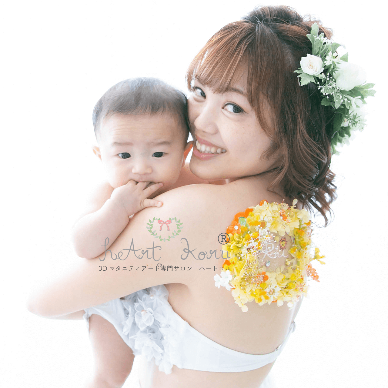 お母さんが赤ちゃんを抱っこしてカメラ目線で撮影している写真。お母さんの肩には、立体のお花のアートである3D授乳ペイントが施されている。