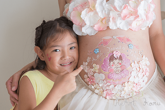 ベーシックコースのベリーペイントのデザイン例。ピンク色のお花がいっぱい散りばめられたマタニティペイント。貝殻の中に天使の女の子の赤ちゃんが描かれていて、子供達の名前が入ったかわいいマタニティアート。子供と一緒に自然光で美しく撮影したマタニティフォト。