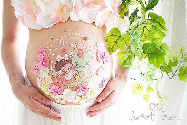 ベーシックコースのベリーペイントのデザイン例。ピンクや白の立体のお花たちがかわいいマタニティペイント。keep smiling（キープスマイリング）のメッセージと、元気いっぱいな恐竜と天使の女の子の赤ちゃんが描かれたおしゃれなマタニティアート。自然光で美しく撮影したマタニティフォト。