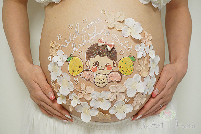 ベーシックコースのベリーペイントのデザイン例。真ん中に、絵の具で天使の女の子赤ちゃんのイラストと、赤ちゃんの両サイドには、ゆずのイラストが入っているとってもかわいいマタニティペイント。赤ちゃんとゆずの周りには、白と淡いサーモンピンクのお花たちがたくさん散りばめられているおしゃれなマタニティアート。上には、白の文字で、welcome baby （ウェルカム ベイビー）と週数が書かれている。お部屋の照明を使って撮影したマタニティフォト。