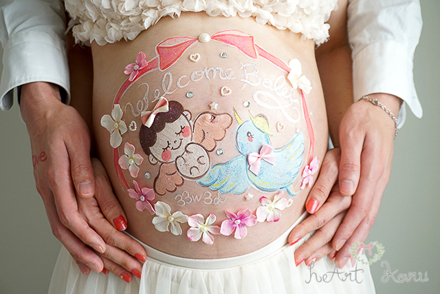 ベーシックコースのベリーペイントのデザイン例。絵の具でピンクのリボンと、天使の女の子赤ちゃんと幸せを運ぶ青い鳥さんのイラストが描かれたかわいいマタニティペイント。周りには、ピンクと白色のお花のデコパーツが散りばめられているおしゃれなマタニティアート。赤ちゃんの上には、白い文字で、welcome baby（ウェルカムベイビー）が書かれている。下には、白い文字で、週数が書かれている。美しい自然光で撮影したマタニティフォト。