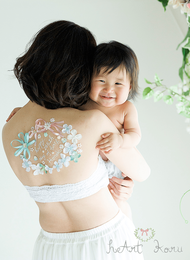 背中に大きく描かれた授乳ペイントは、水色のリボンとピンクのリボンが印象的です。タンポポのお花もワンポイントで添えられています。赤ちゃんを抱きかかえたシーンを撮影した授乳フォトです。