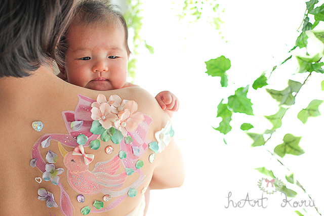 背中にコウノトリが描かれた授乳ペイント。コウノトリ全体を紫のリボンがおおい、緑の花びらがおしゃれさを演出しているデザインです。赤ちゃんを抱きかかえているシーンを撮影した1枚です。