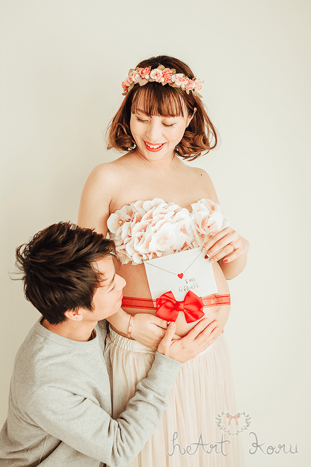 マタニティフォト（ペイント前）。小花の冠と、薄ピンクのブラとふんわりロングスカートが可愛いマタニティフォトの衣装。妊婦さんのお腹に赤いリボンが巻かれ、そこに赤ちゃんへのラブレターを挟み、パパが寄り添うように写っているお洒落なマタニティ写真。
