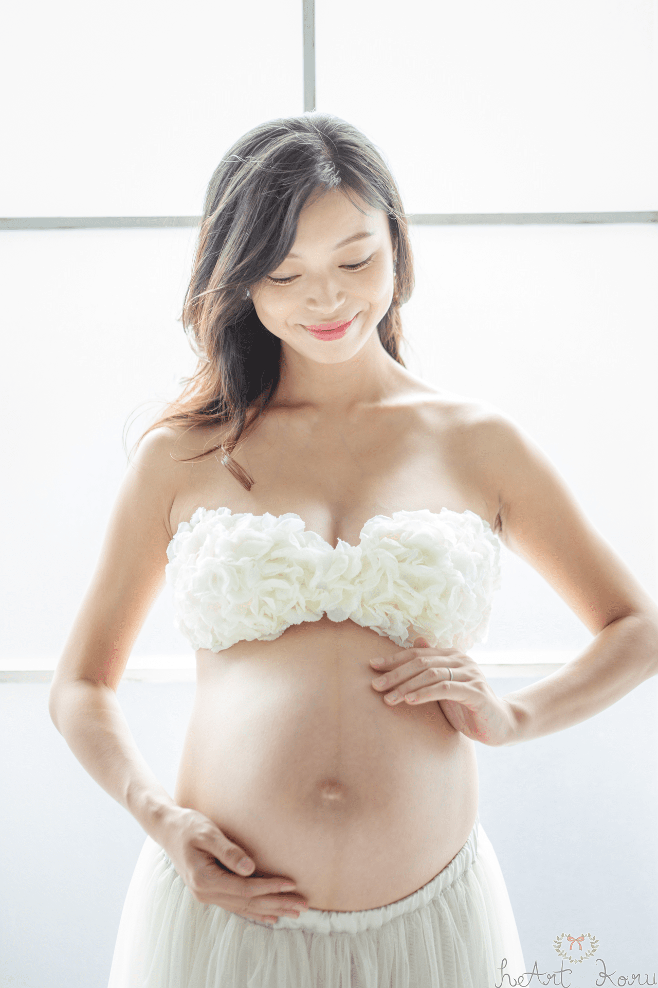 マタニティフォト（ペイント前）。妊婦さんは、ホワイトのブラトップと、ロングスカートのマタニティフォトの衣装を着ている。妊婦さんが、優しく両手をお腹の赤ちゃんを守るように手を添えているマタニティ写真。美しい自然光マタニティ撮影。