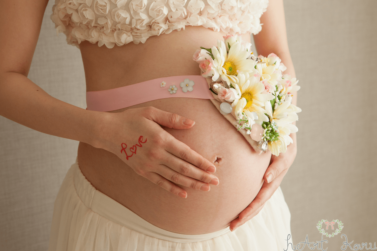 マタニティフォト（ペイント前）。妊婦さんがお花のマタニティサッシュをお腹につけてポーズをとっているおしゃれなマタニティ撮影。マタニティフォトの衣装は、ホワイトのブラトップとふんわりロングスカート。自然光が綺麗なマタニティ写真。