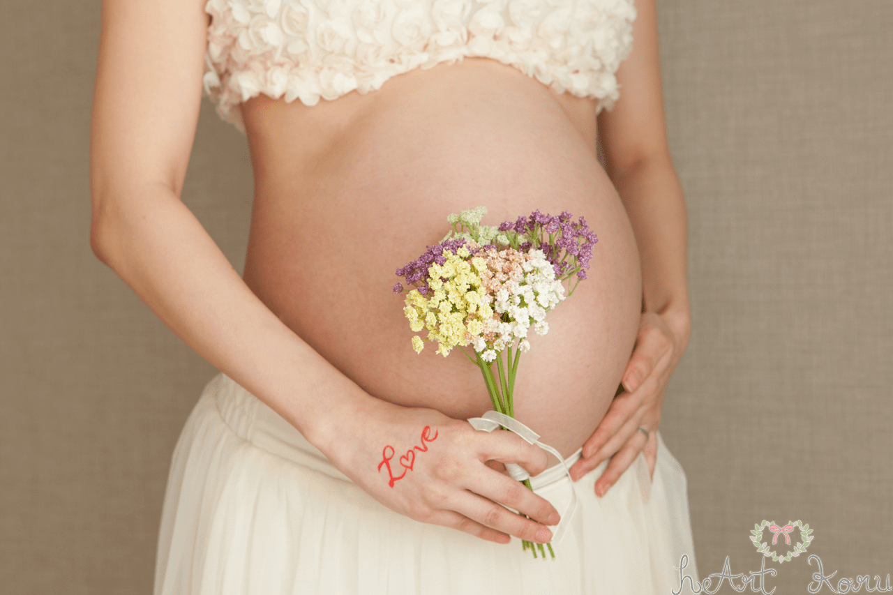 マタニティフォト（ペイント前）。妊婦さんがお腹に、お花のブーケを置いてポーズをとっているおしゃれなマタニティ撮影。マタニティフォトの衣装は、ホワイトのブラトップとふんわりロングスカート。自然光が綺麗なマタニティ写真。