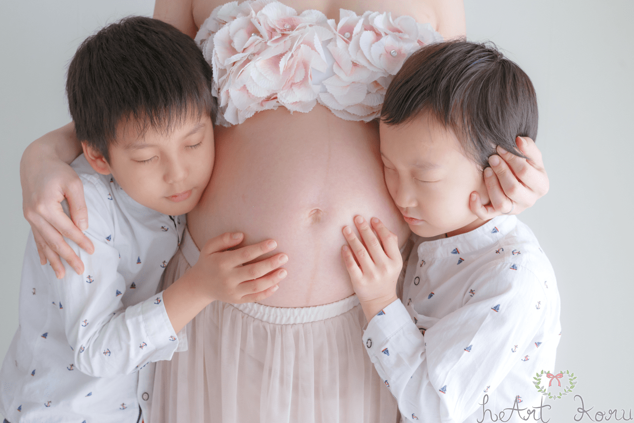マタニティフォト（ペイント前）。妊婦さんのお腹の両サイドにお兄ちゃんが抱きついて写っているマタニティ写真。マタニティフォトの衣装は、お花の冠、薄ピンクのブラトップとふんわりロングスカート。自然光が綺麗なマタニティ写真。