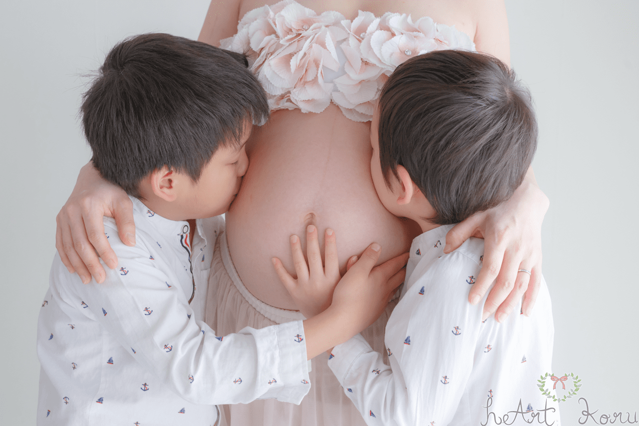 マタニティフォト（ペイント前）。妊婦さんのお腹の両サイドにお兄ちゃんがキスして写っているマタニティ写真。マタニティフォトの衣装は、お花の冠、薄ピンクのブラトップとふんわりロングスカート。自然光が綺麗なマタニティ写真。