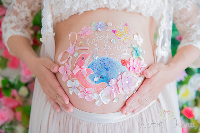 お姫様コースのベリーペイントのデザイン例。白とピンクとブルーのお花で彩られているとてもおしゃれなマタニティペイント。妊娠週数とhappy maternityのメッセージが入った、ピンクイルカと黄色とピンクの蝶々のイラストが描かれているマタニティアート。自然光で撮影したマタニティフォト。