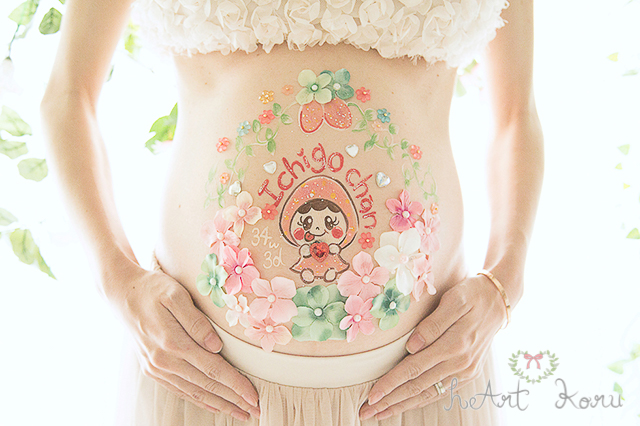 お姫様コースのベリーペイントのデザイン例。緑とピンクのお花で彩られているとてもおしゃれなマタニティペイント。赤ちゃんの名前である、「Ichigo-chan」（イチゴちゃん）とメッセージが入っている。赤ちゃんがいちごの着ぐるみを着ているかわいいイラストが描かれているマタニティアート。自然光で撮影したマタニティフォト。