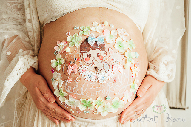 お姫様コースのベリーペイントのデザイン例。緑とピンクのデコパーツのお花で彩られているとてもおしゃれなマタニティペイント。妊娠週数とwelcome baby （ウェルカムベイビー）のメッセージが入っている。二匹の犬と赤ちゃんのイラストが描かれている可愛いマタニティアート。自然光で撮影したマタニティフォト。