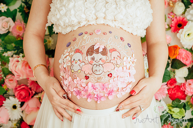 お姫様コースのベリーペイントのデザイン例。白とピンクのデコパーツのお花で彩られているとてもおしゃれなマタニティペイント。妊娠週数とwelcome baby （ウェルカムベイビー）のメッセージが入っている。赤ちゃんと、両サイドには、二羽の鳥さんのイラストが描かれているとってもかわいいマタニティアート。フラワーウォールで撮影したおしゃれでかわいいマタニティフォト。