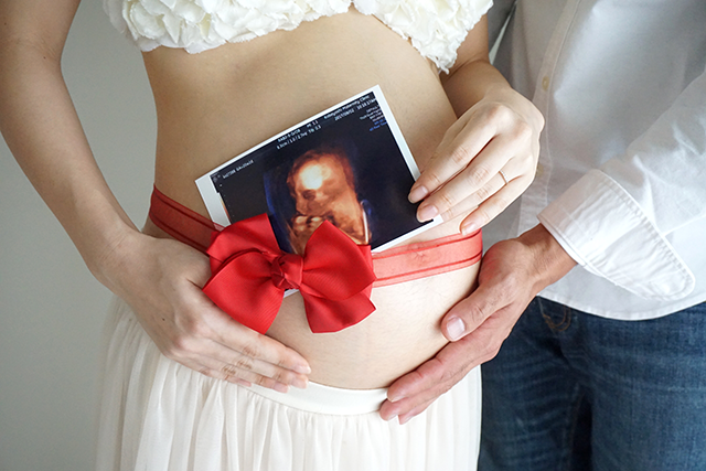 ベリーペイント（マタニティペイント）の前の、マタニティフォト。妊婦さんのお腹に、赤いリボンが巻かれ、そこに赤ちゃんにエコー写真が添えられている。2人の手が優しくお腹を包み込んでる。